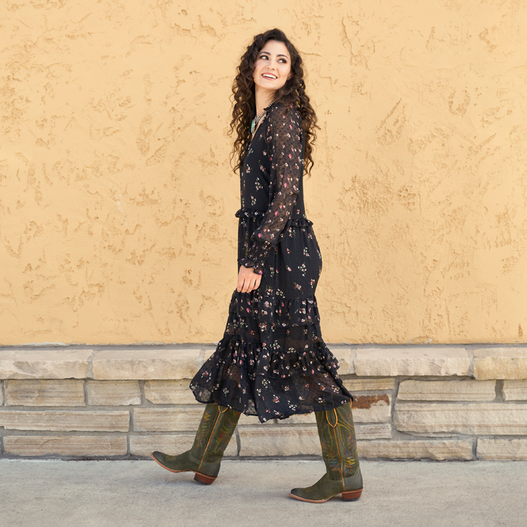 A woman with brown curly hair wearing green Justin boots walking down a sidewalk and looking back away from the camera.
